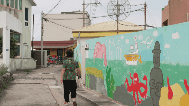 Back of elderly woman walking past colorful mural on village street.