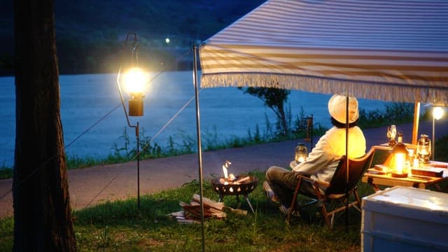 Person relaxing under tent canopy in a campsite beside a river in the evening