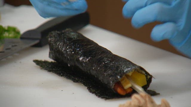 Hands wearing gloves preparing a kimbap rollp on cutting board