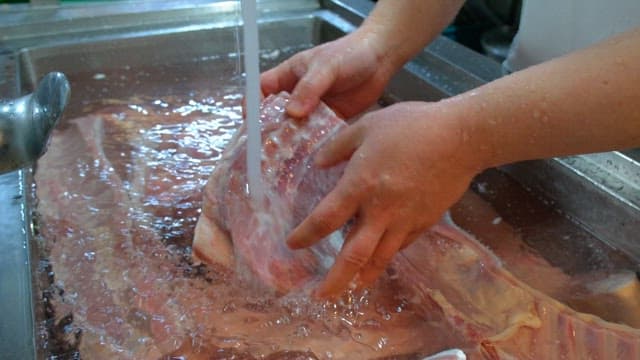 Washing large cuts of raw meat under running water in a kitchen