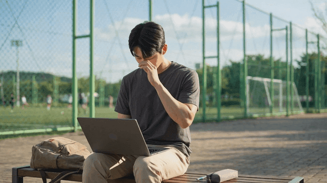 A young man working on a laptop by a sports field