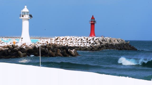 Snow-covered beach with crashing waves and a lighthouse on a breakwater