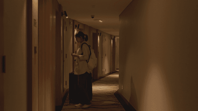 Woman walking down a hotel hallway with her suitcase into her room.