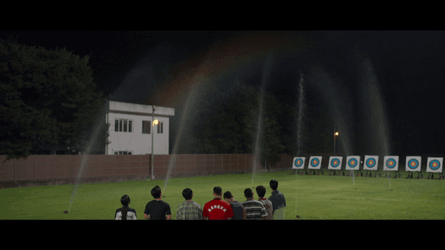Back view of people observing archery practice range with sprinklers turned on at night