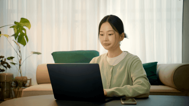 Woman drinking coffee and looking at her phone while working on her laptop at home