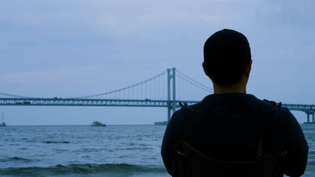 Man Sitting on a Chair Looking at the Sea and Bridge