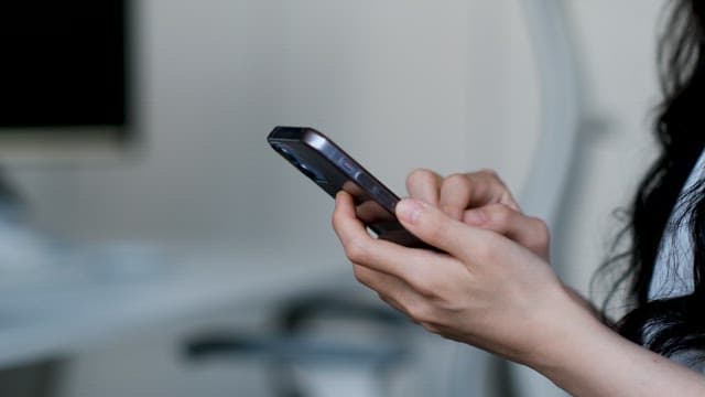 하얀 방 안 책상 앞에서 핸드폰을 사용하는 검은 긴 머리의 여자