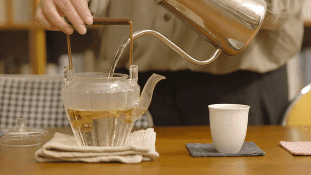 Person pouring hot water into glass teapot containing tea leaves