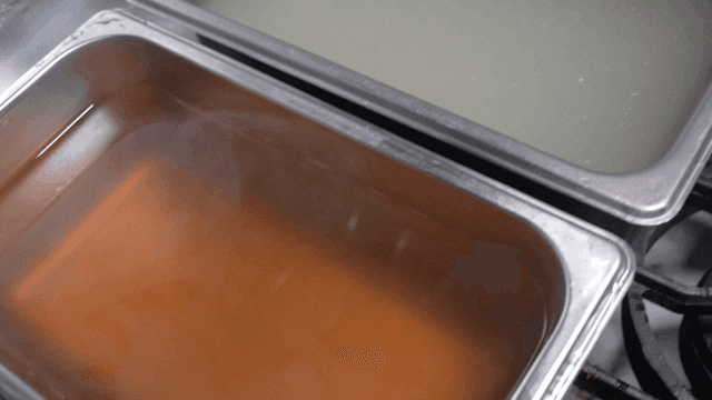 Two trays of broth simmering on a stove
