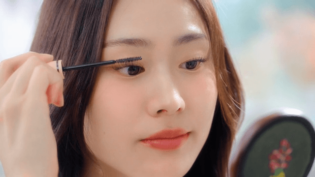 Woman carefully applying mascara to her lashes with a mirror indoors
