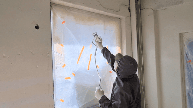 Worker spraying paint with spray gun on wall near window