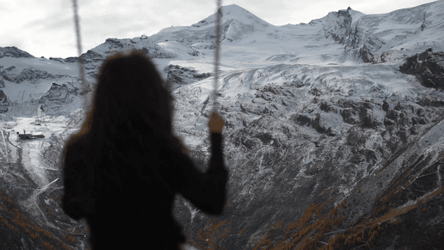 Back view of woman riding swing with snow-covered mountains in background