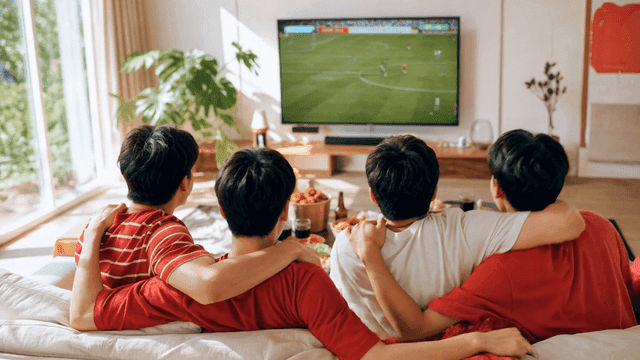 Friends watching soccer match with their arms around each other's shoulders on sofa at home