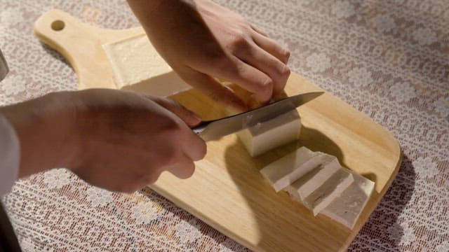 Person Cutting Tofu on a Wooden Board with a Knife