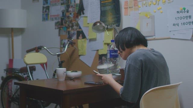 어수선한 방의 식탁에 앉아 핸드폰을 만지는 여자