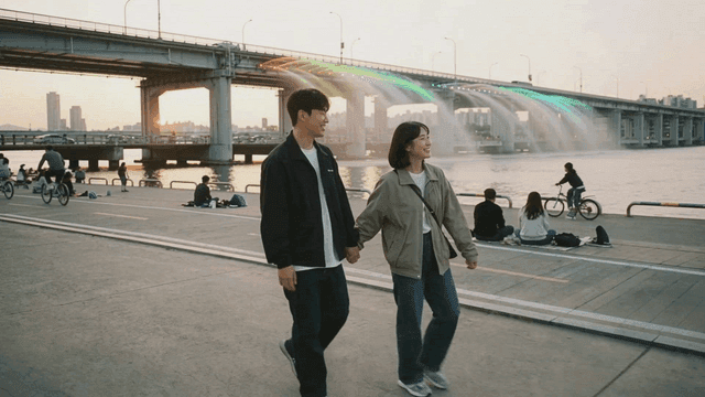 Couple walking hand in hand along riverside next to bridge with fountain