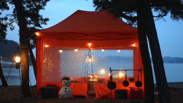 Snowman outside a lit orange tent near a tranquil sea at dusk