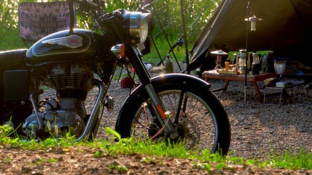 Motorcycle parked near a campsite in a forest with camping gear and tents set up under morning sunli