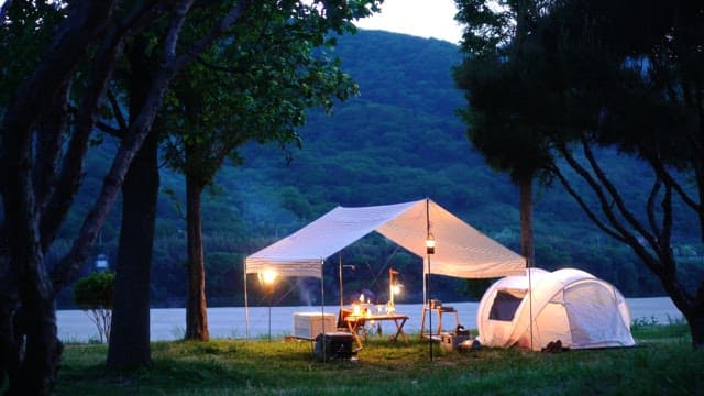 Person relaxing under tent canopy in a campsite beside a river in the evening