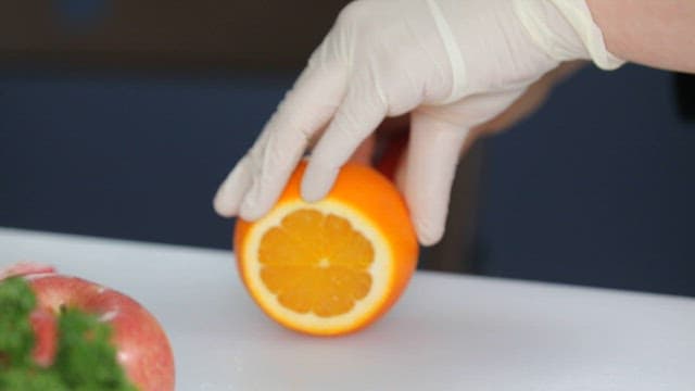 Gloved hand slicing an orange on a cutting board