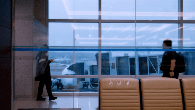 Passengers walking in airport terminal where airplanes are visible