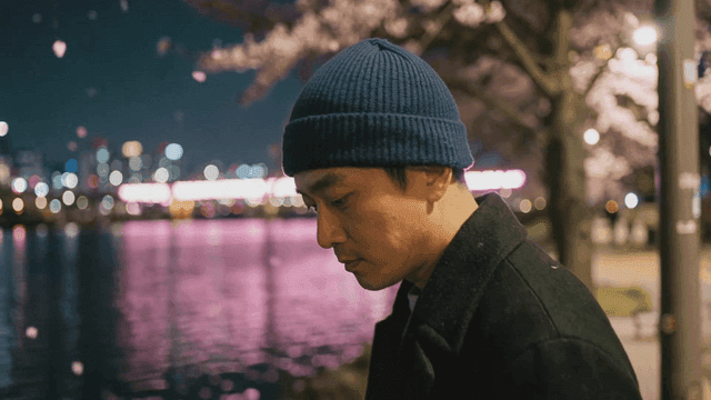 Man wearing beanie by river where cherry blossoms are falling at night