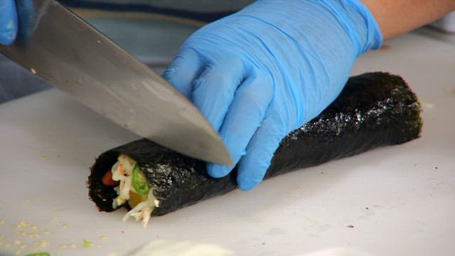 Hands wearing gloves cutting a kimbap rollp on cutting board