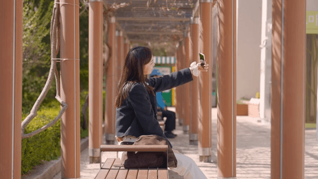 Woman sitting on park bench taking selfie while waiting for someone