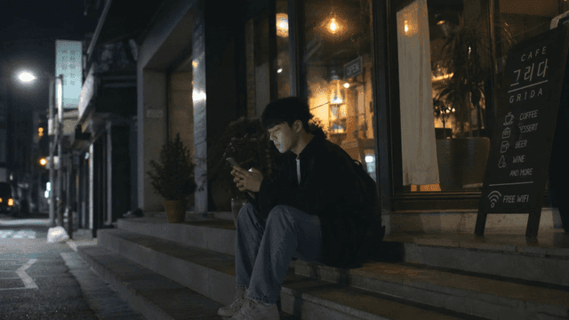 Young man sitting outside cafe at night looking at his phone