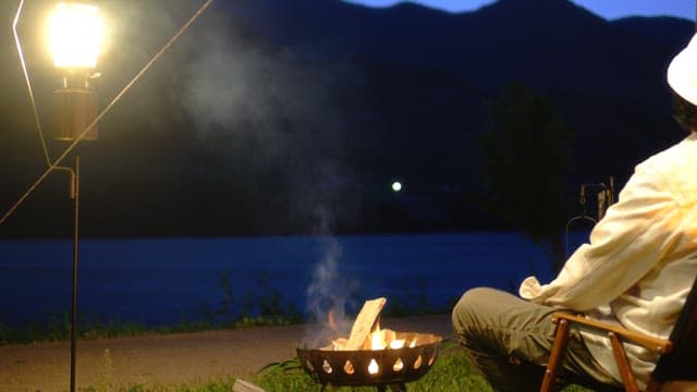 Evening camping by the serene riverside with a person sitting beside a campfire