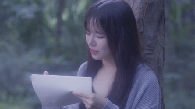 숲 속 나무에 기대어 종이에 글을 쓰는 머리 긴 여자