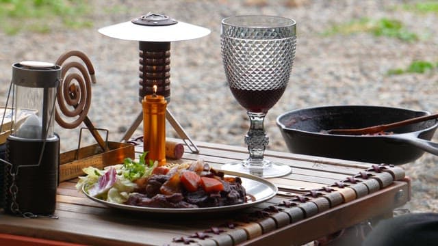 Outdoor meal setup with red wine, cooked meat and camping gear