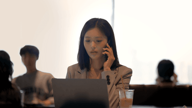Female office worker talking on phone while looking at her laptop in cafe