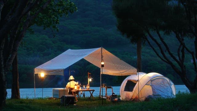 Person relaxing under tent canopy in a campsite beside a lake in the evening