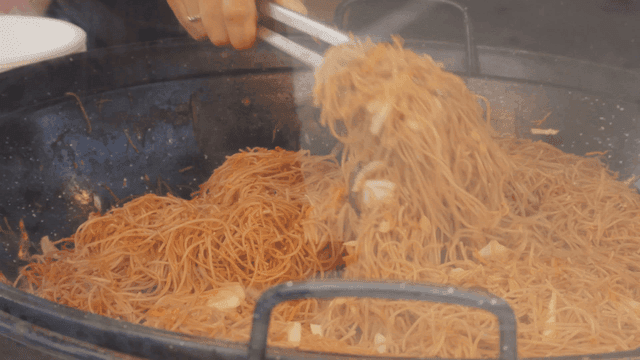 Tongs for stir-frying noodles in large pan and placing them in bowl