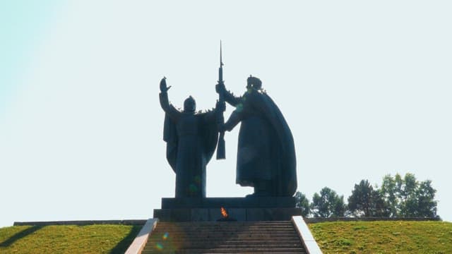 Statues of two figures holding a spear with eternal flame at the base in a park.