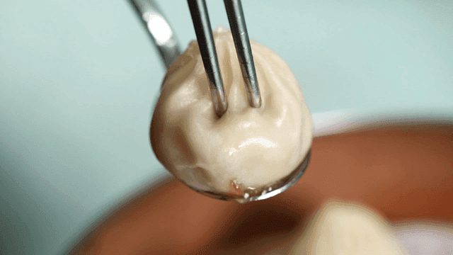 Chopsticks pressing down on dumpling on spoon with flowing broth