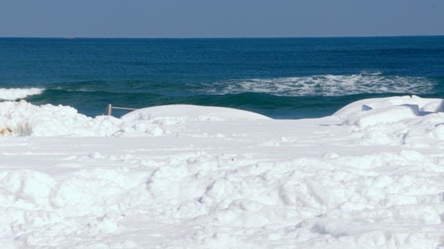 Ocean waves crashing onto a snow-covered beach under a clear blue sky