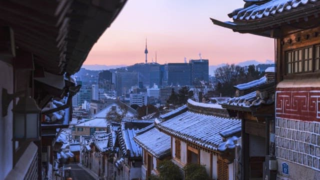 Scenic view of a traditional Korean village with snowy rooftops during sunrise, overlooking city sky