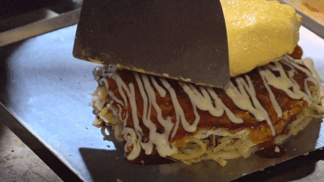 Egg omelette placed on okonomiyaki on griddle by spatula