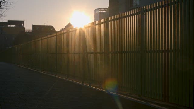 해질녘 건물을 둘러싼 높은 펜스 앞을 따라 걷는 남자