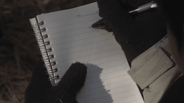 장갑 낀 손이 야외에서 노트에 글을 쓰는 모습