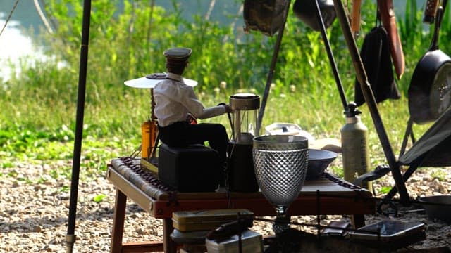 Outdoor camping setup with a puppet cooking food on a table by the riverside.