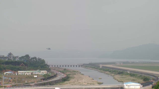 Helicopter flying over a river near countryside buildings and a runway