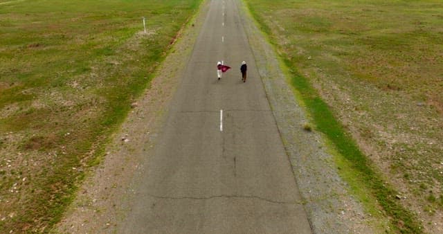 Two people walking on a long road surrounded by wide grasslands