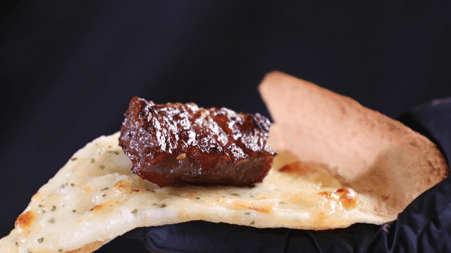 Glovenly hand holding grilled beef short ribs on cheese pizza