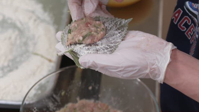 Gloved hands stuffing minced meat between perilla leaves