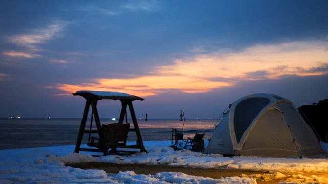 Tent and a wooden swing near the sea at sunset with a snowy landscape