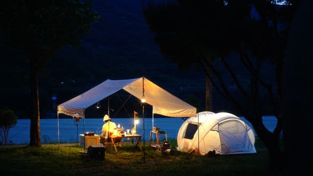 Person relaxing under tent canopy in a campsite beside a river in the night