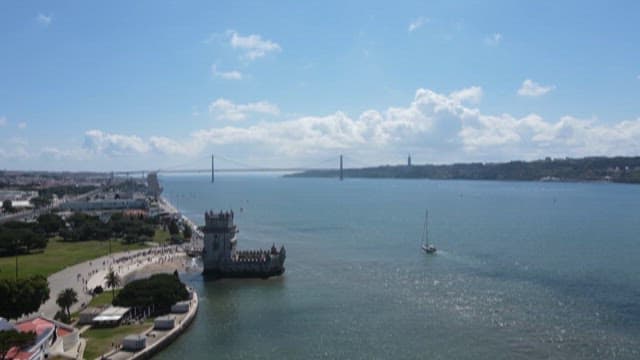 Clear Day View of Belém Tower by the Water with Tourists and Suspension Bridge in the Distance